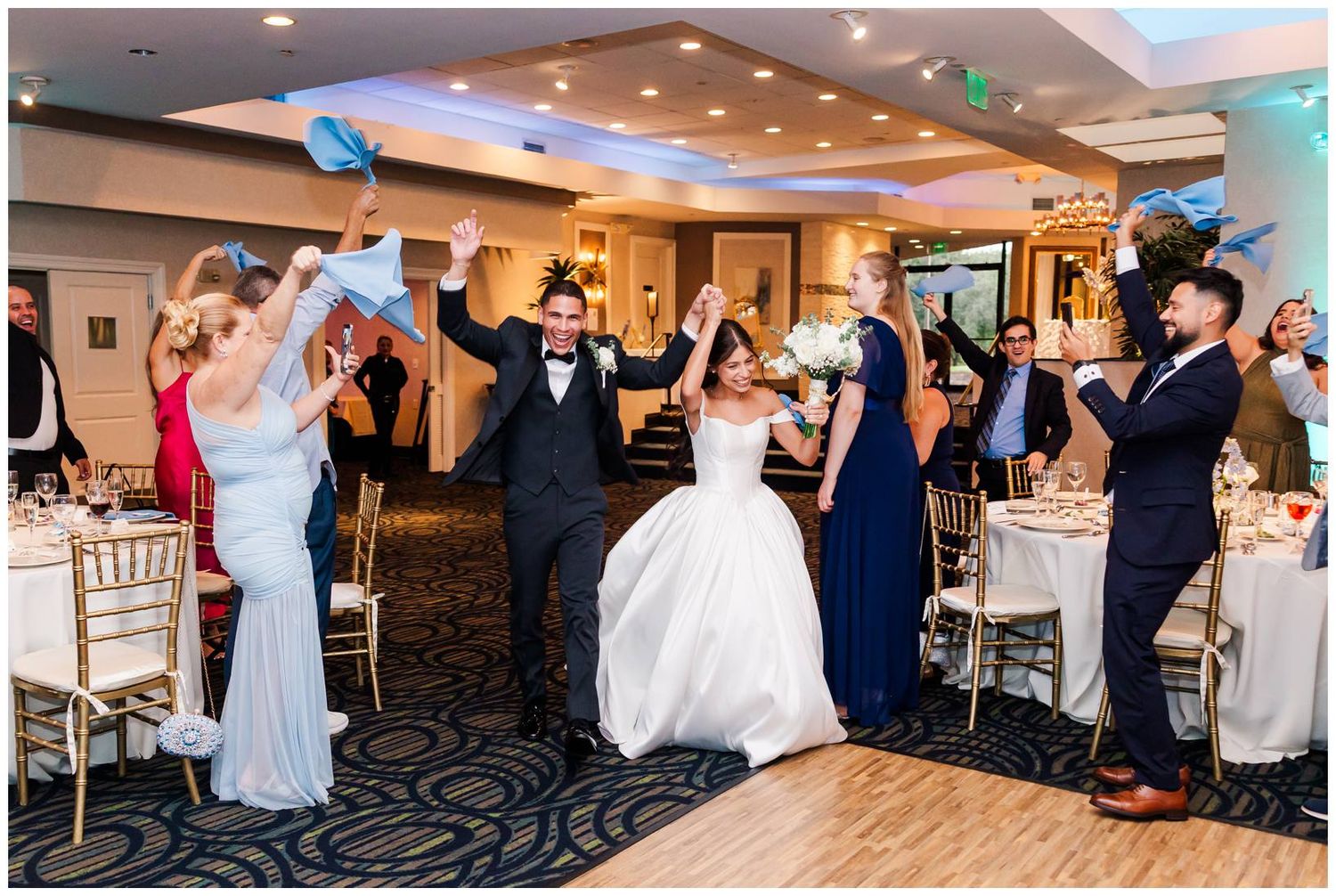 Bride and groom make grand entrance into wedding reception as guests cheer and wave blue paper fans in celebration.