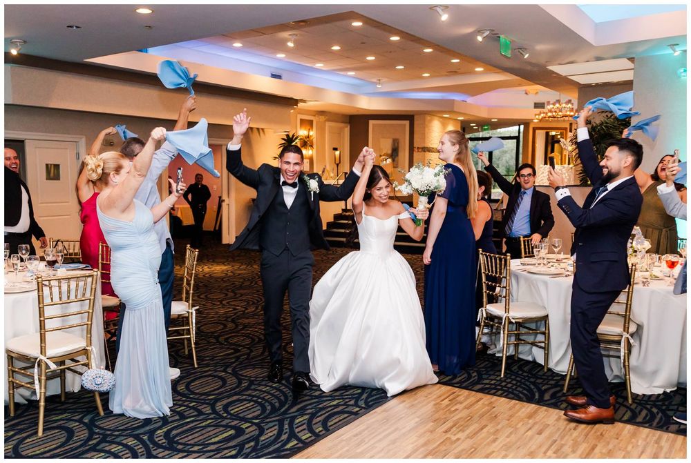 Bride and groom make grand entrance into wedding reception as guests cheer and wave blue paper fans in celebration.