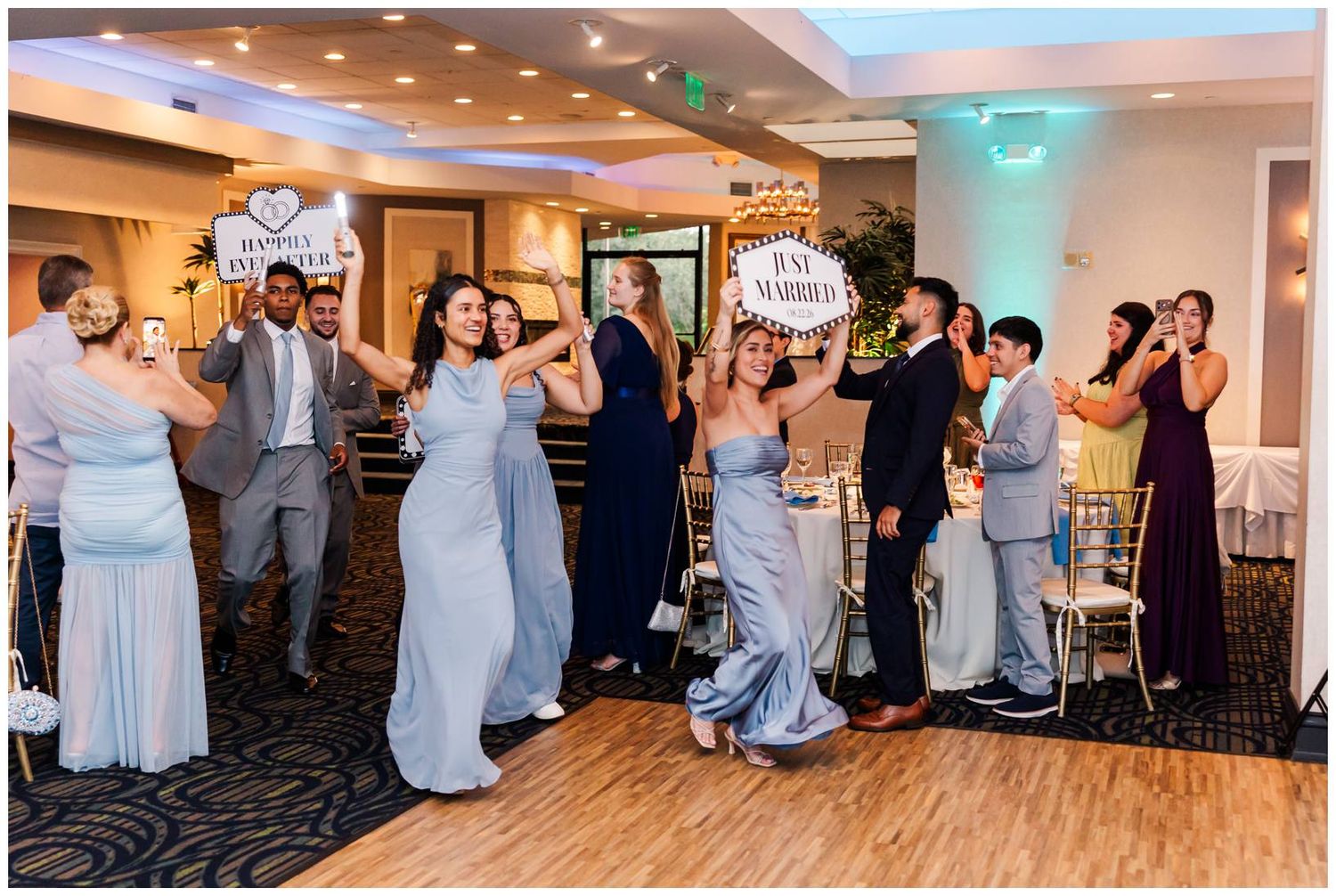 Wedding party celebrating at reception, holding just married signs while dancing in elegant ballroom venue.