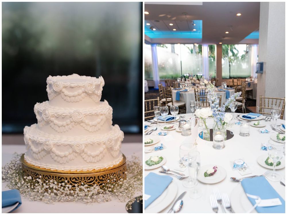 Elegant three-tier white wedding cake on gold stand beside beautifully set reception tables with blue accents.