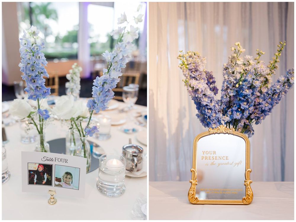 Elegant wedding reception centerpieces featuring blue and purple delphinium flowers in glass vases on white tablecloths.
