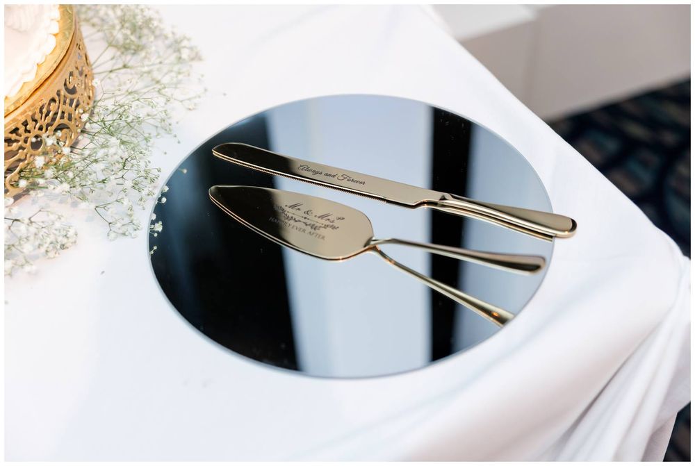 Gold wedding cake server and knife set displayed on a round mirror tray with white floral decor.