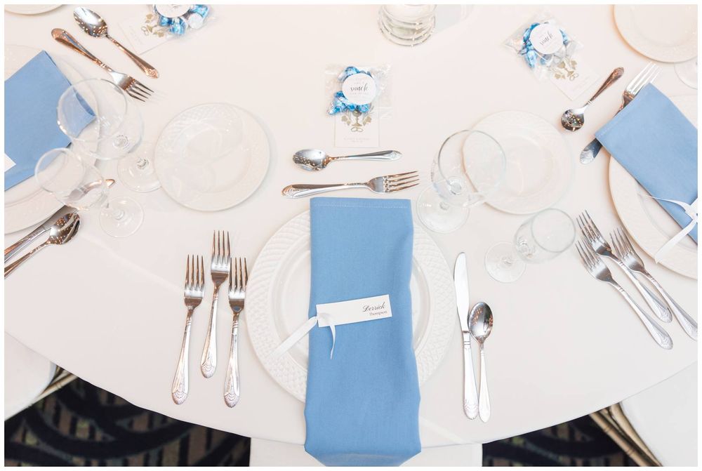 Elegant wedding reception table setting with blue napkins, silver cutlery, and white plates on a white tablecloth.