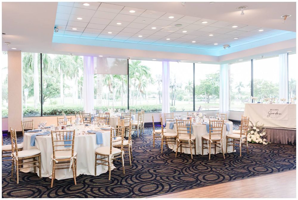 Elegant wedding reception venue with gold chiavari chairs, round tables, and large windows overlooking palm trees.