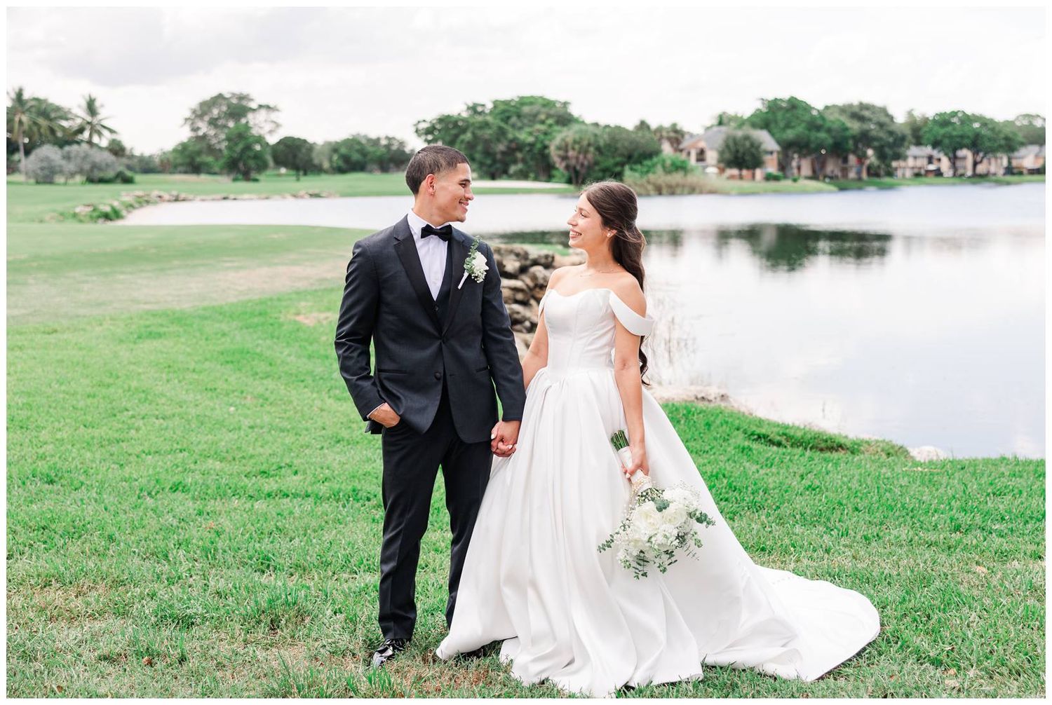 Bride in an elegant off-shoulder gown and groom in a black tuxedo share a loving gaze near a peaceful lake.