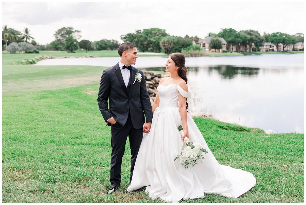 Bride in an elegant off-shoulder gown and groom in a black tuxedo share a loving gaze near a peaceful lake.