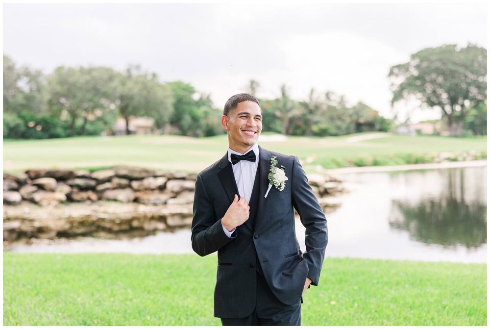 Dapper groom in a black tuxedo smiling confidently outdoors near a scenic pond and lush greenery.