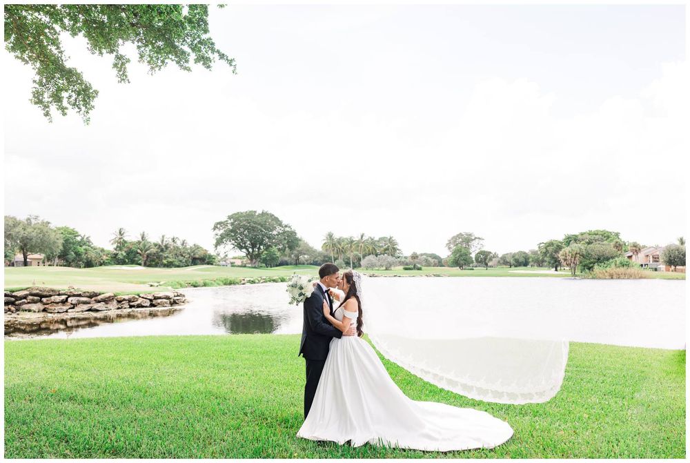 Bride and groom share a romantic embrace on a green lawn with a serene lake and trees in the background.