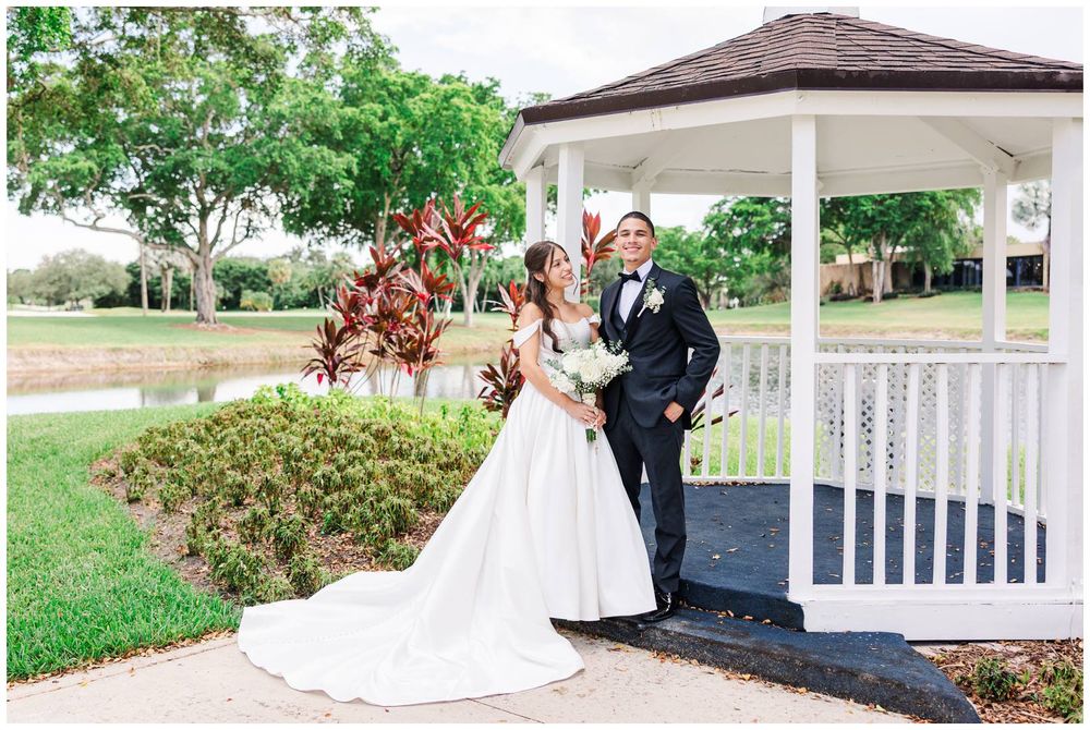 Bride and groom posing together by white gazebo near pond in outdoor wedding venue setting.