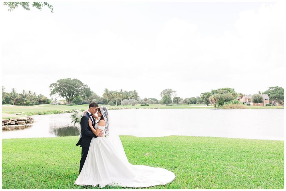Bride and groom share a romantic embrace on a green lawn with a tranquil pond backdrop.