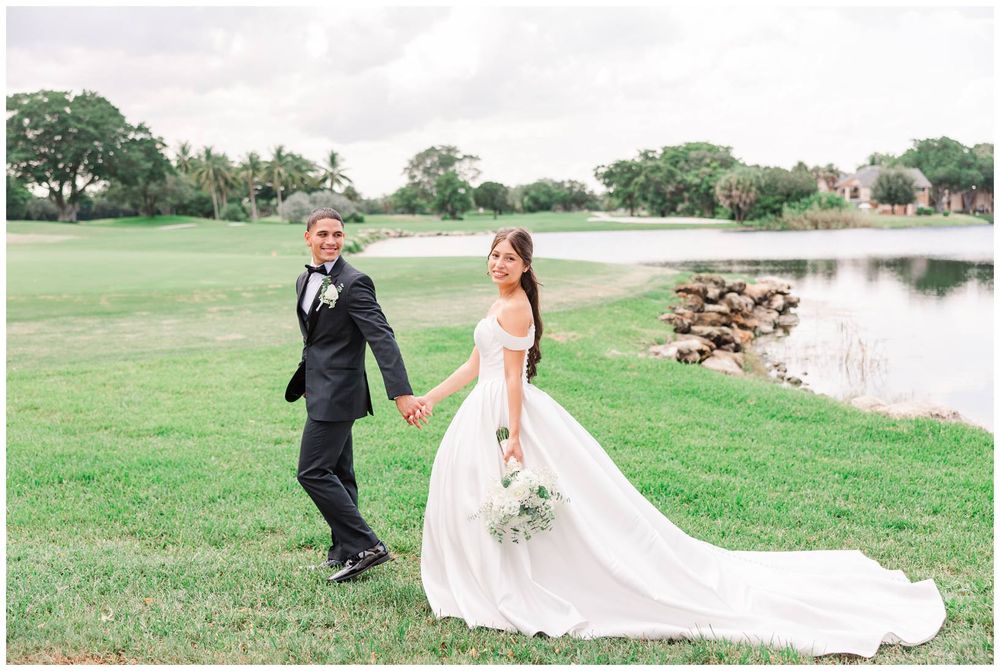 Bride and groom holding hands walking on lush green golf course lawn near pond on their wedding day.