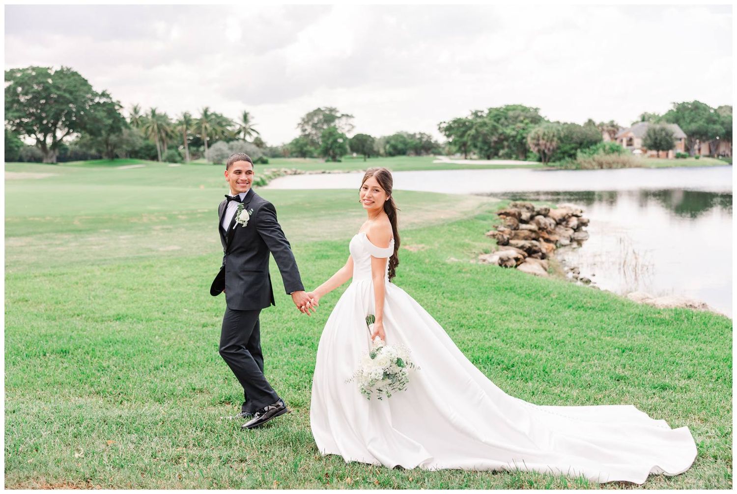 Bride and groom holding hands walking on lush green golf course lawn near pond on their wedding day.