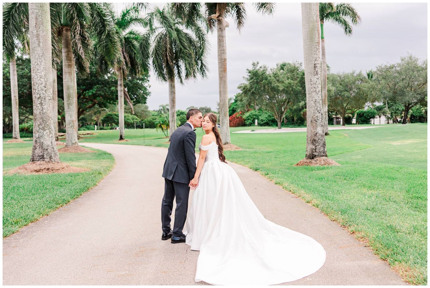 Bride and groom share a romantic kiss on a winding path surrounded by tall palm trees at an outdoor wedding venue.