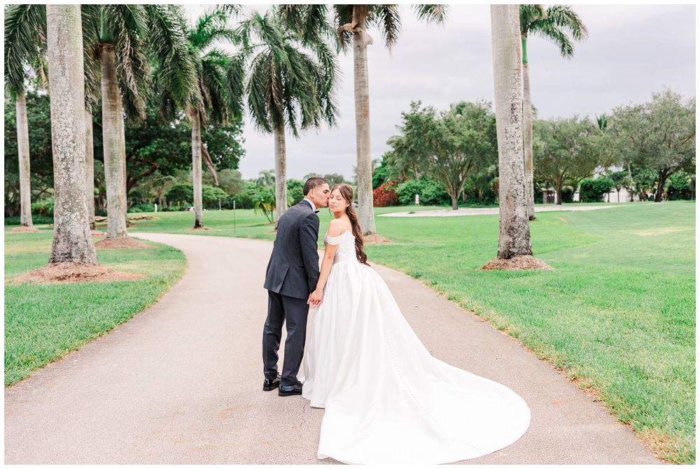 Bride and groom share a romantic kiss on a winding path surrounded by tall palm trees at an outdoor wedding venue.