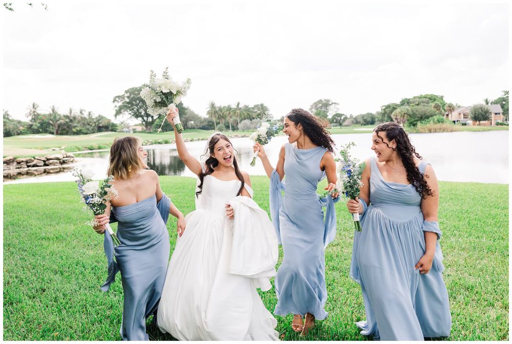 Bride in white gown celebrating with bridesmaids in dusty blue dresses holding bouquets on green lawn.