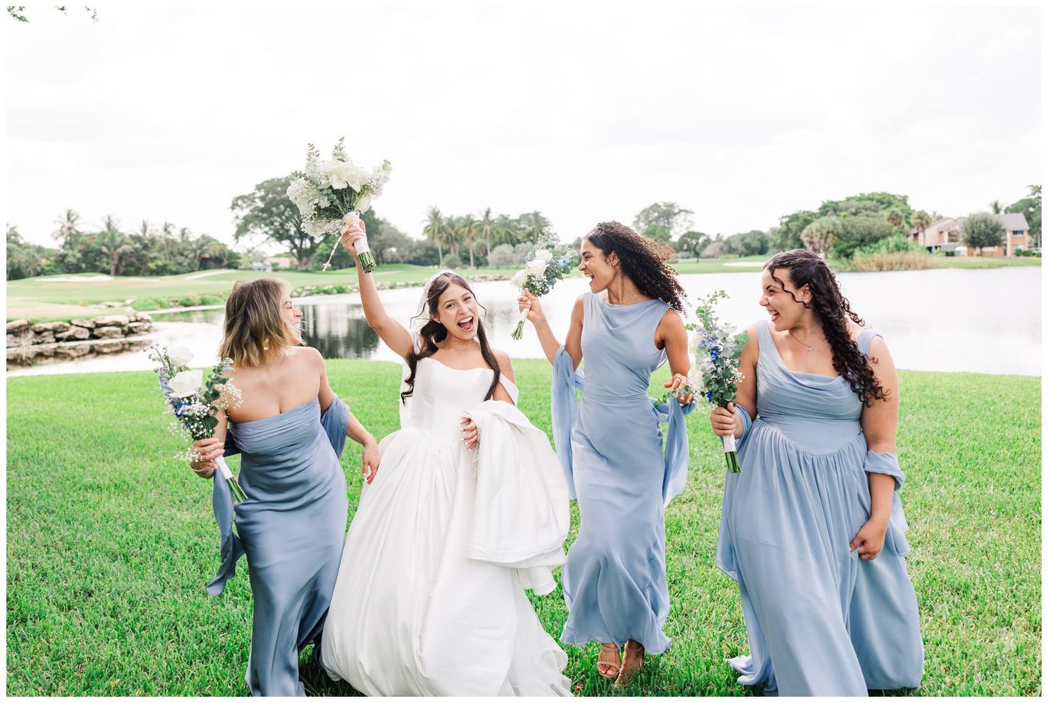 Bride in white gown celebrating with bridesmaids in dusty blue dresses holding bouquets on green lawn.
