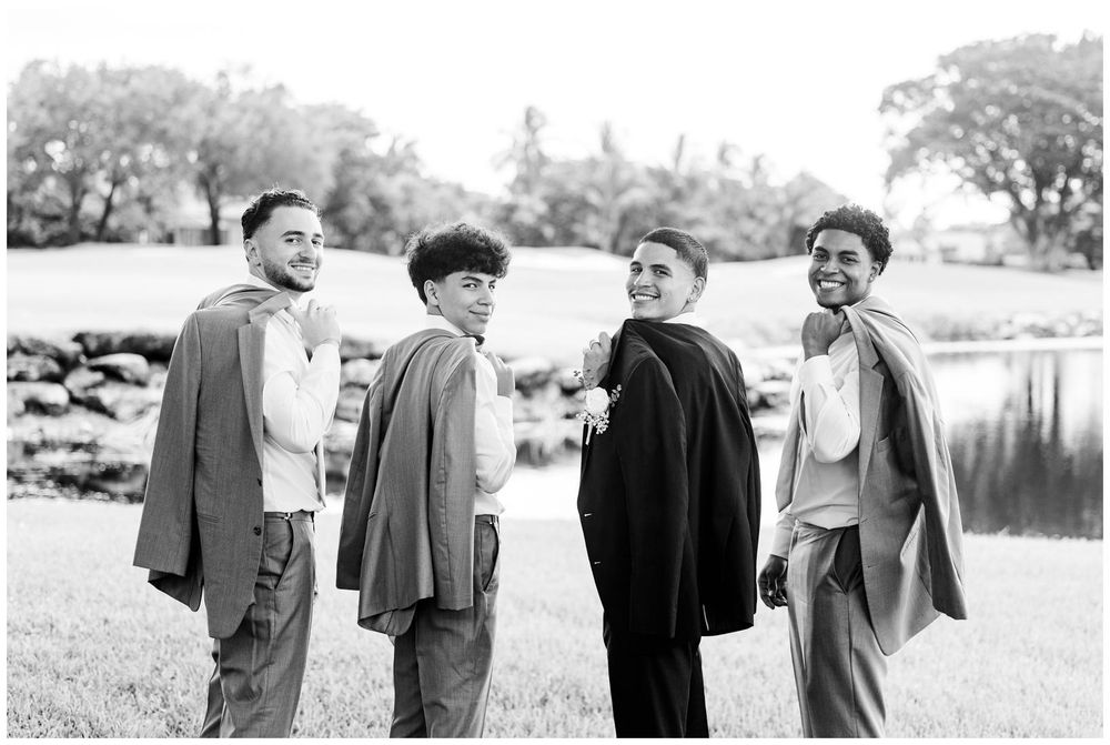 Four groomsmen in suits laughing outdoors near a pond in a black and white wedding photo.