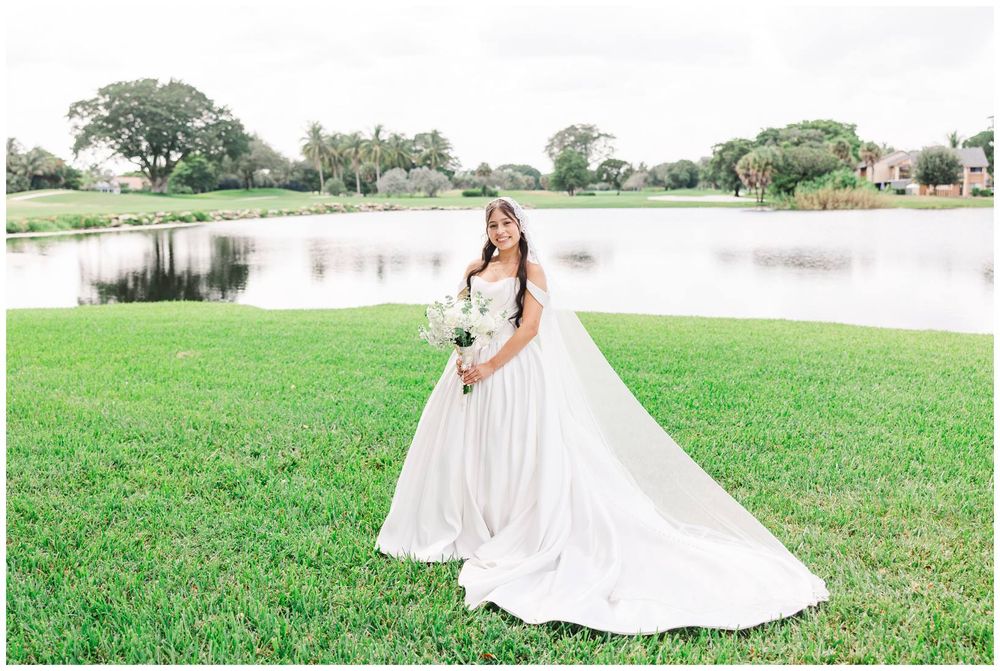 Bride in flowing white gown with long veil holding bouquet on green lawn beside serene pond.