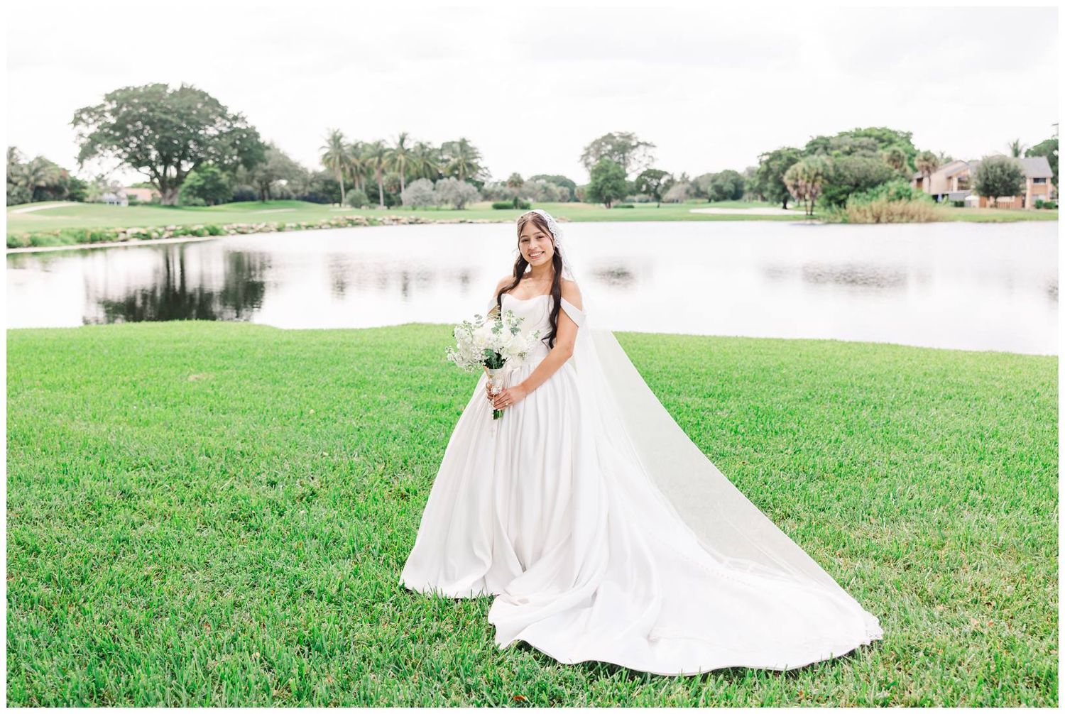 Bride in flowing white gown with long veil holding bouquet on green lawn beside serene pond.