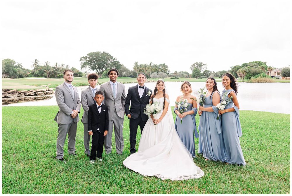 Wedding party in blue and gray attire poses with bride and groom on a green lawn near a pond.