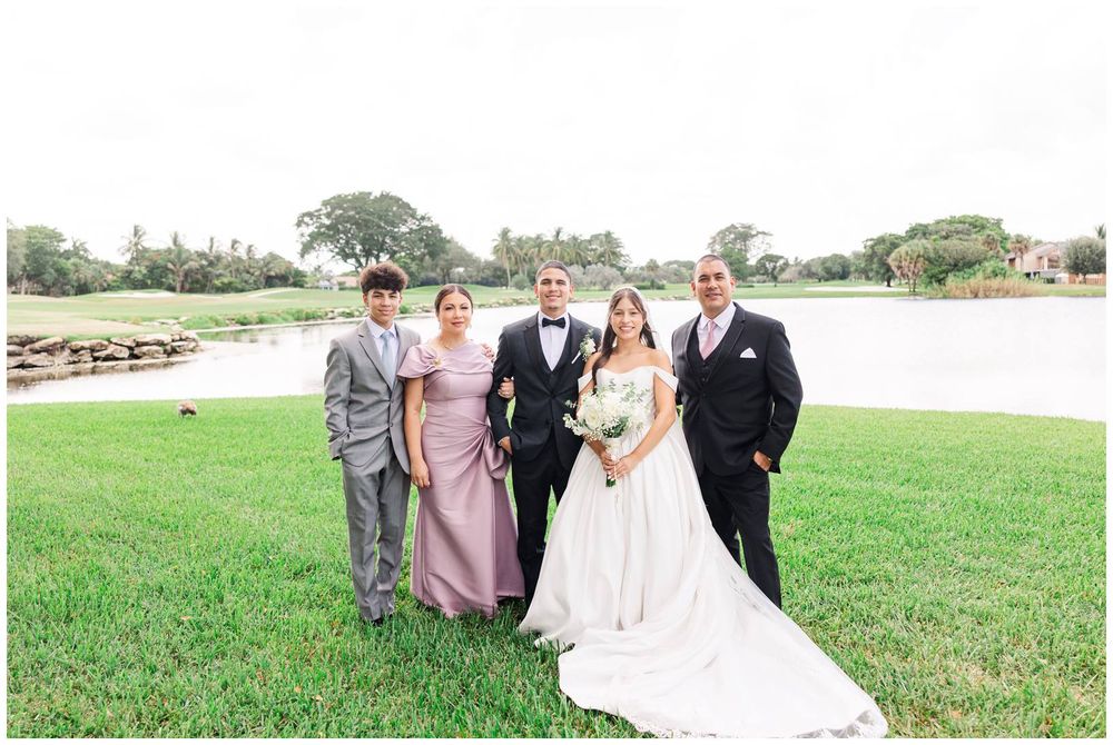 Wedding party family portrait with bride and groom posing beside pond on green grass.