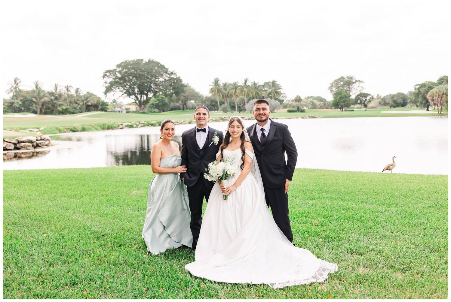 Bride and groom pose with wedding party guests on lush green golf course lawn near a serene pond.