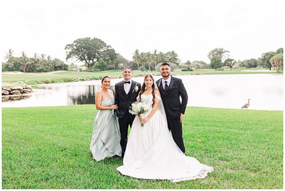 Bride and groom pose with wedding party guests on lush green golf course lawn near a serene pond.