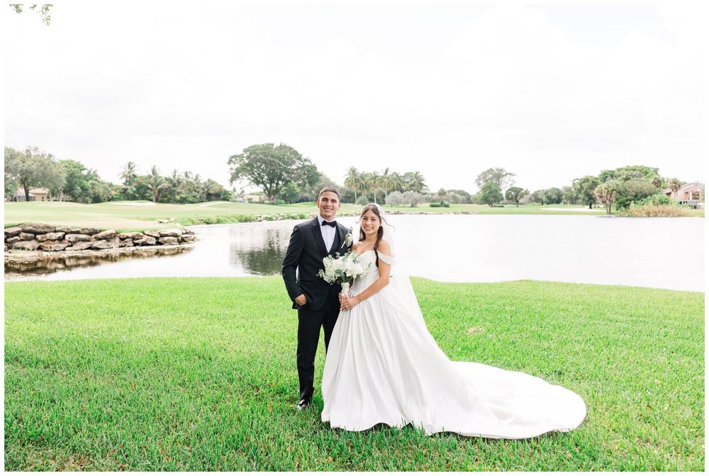 Bride and groom pose together on a lush green lawn beside a serene pond on their wedding day.