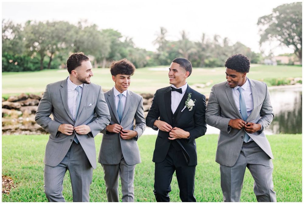 Groom in black tuxedo laughing with three groomsmen in gray suits on green lawn.