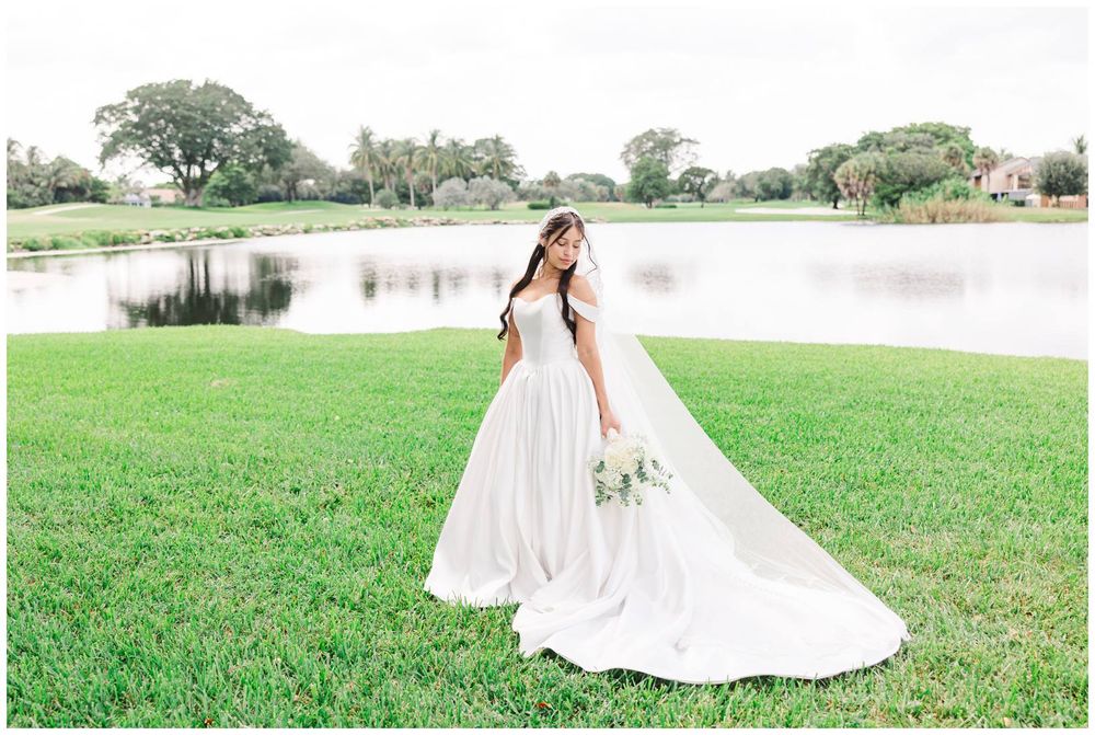 Bride in elegant white ballgown with long veil holding bouquet beside a serene pond.