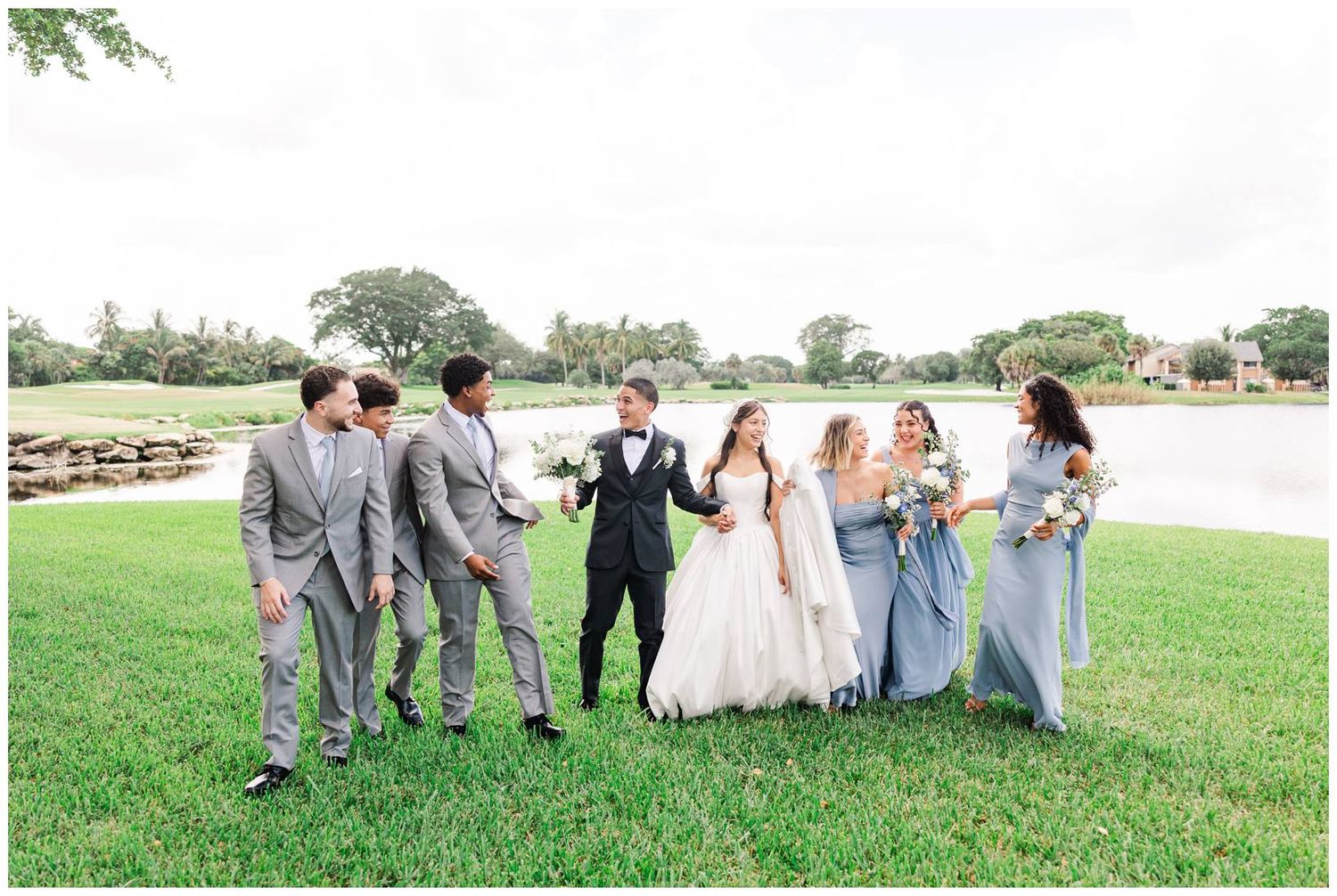 Wedding party walks on green lawn near a lake, bride and groom centered with groomsmen and bridesmaids in blue-gray attire.