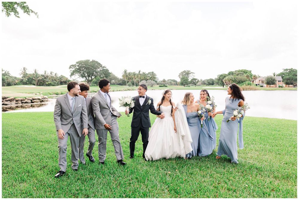 Wedding party walks on green lawn near a lake, bride and groom centered with groomsmen and bridesmaids in blue-gray attire.