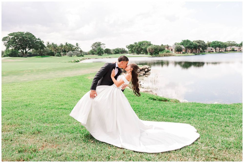 Groom dips bride in white gown for a romantic kiss by a serene pond on a lush green golf course.
