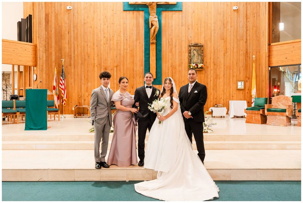 Bride in elegant ball gown poses with groom and family members inside a warmly lit Catholic church.