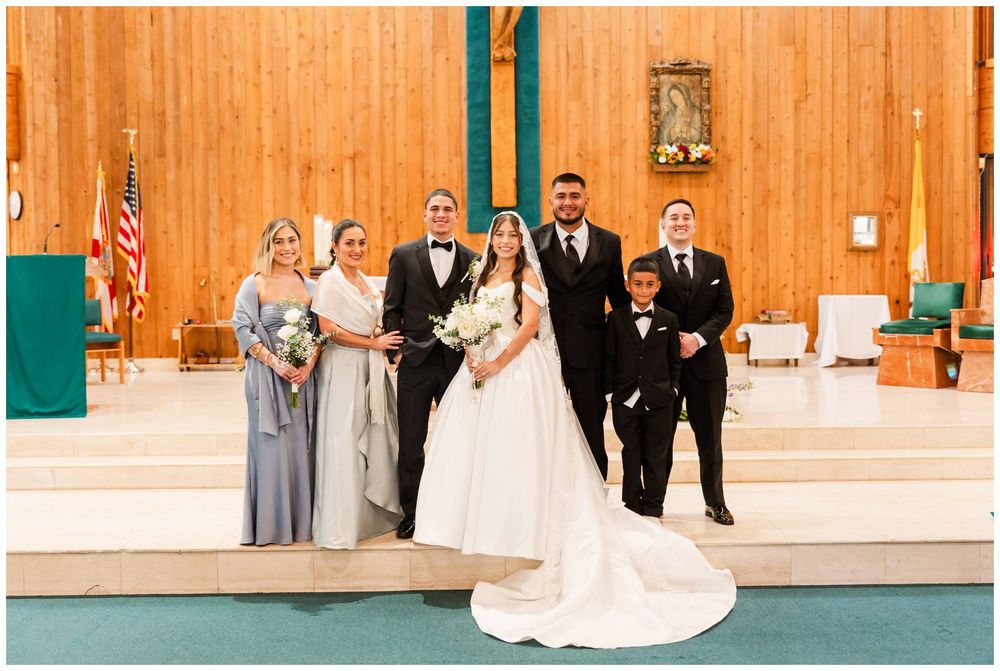 Wedding party poses with bride and groom inside a Catholic church with wooden walls and teal accents.