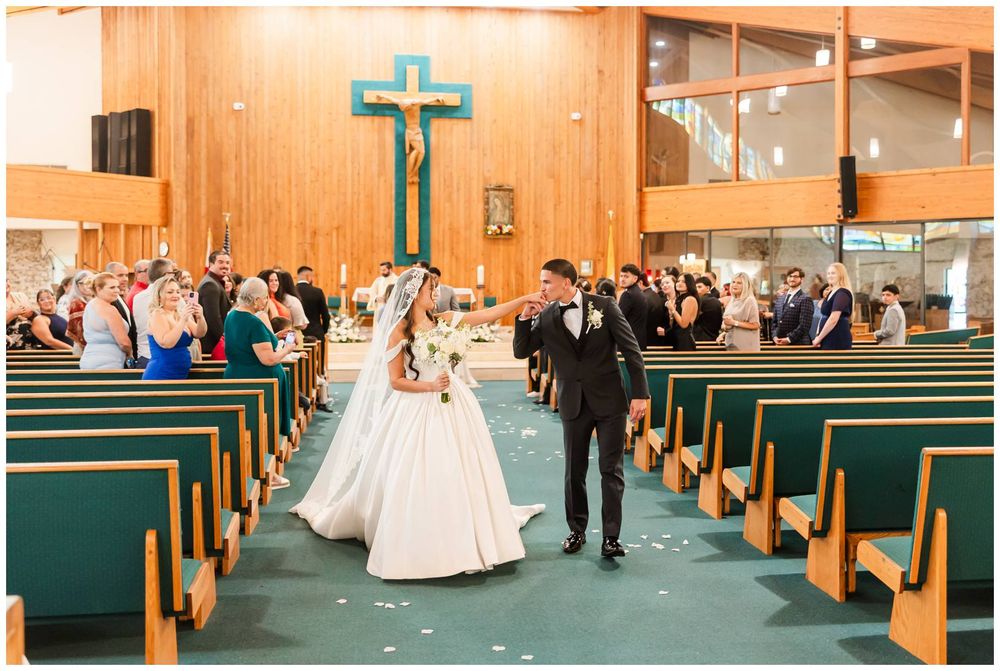 Bride and groom walk down church aisle after wedding ceremony, surrounded by guests in wooden pews beneath a teal cross.