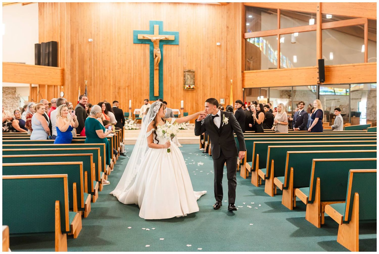 Bride and groom walk down church aisle after wedding ceremony, surrounded by guests in wooden pews beneath a teal cross.