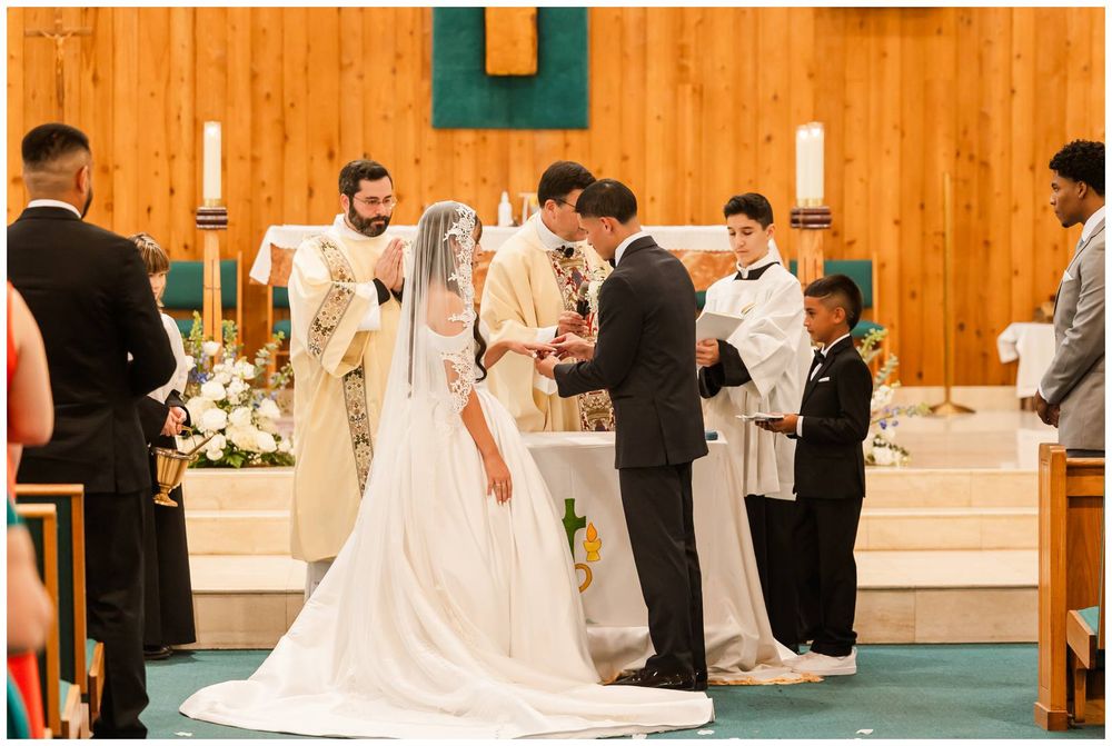 Couple exchanging rings at church altar with priest and wedding party during ceremony.
