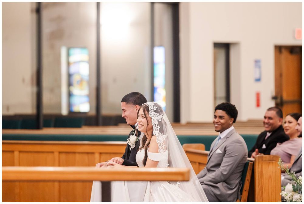 Bride and groom kneel at church altar during wedding ceremony, with guests seated in wooden pews behind them.