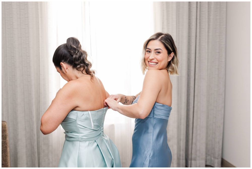 Smiling bridesmaid in blue strapless dress helping another bridesmaid zip up her gown.