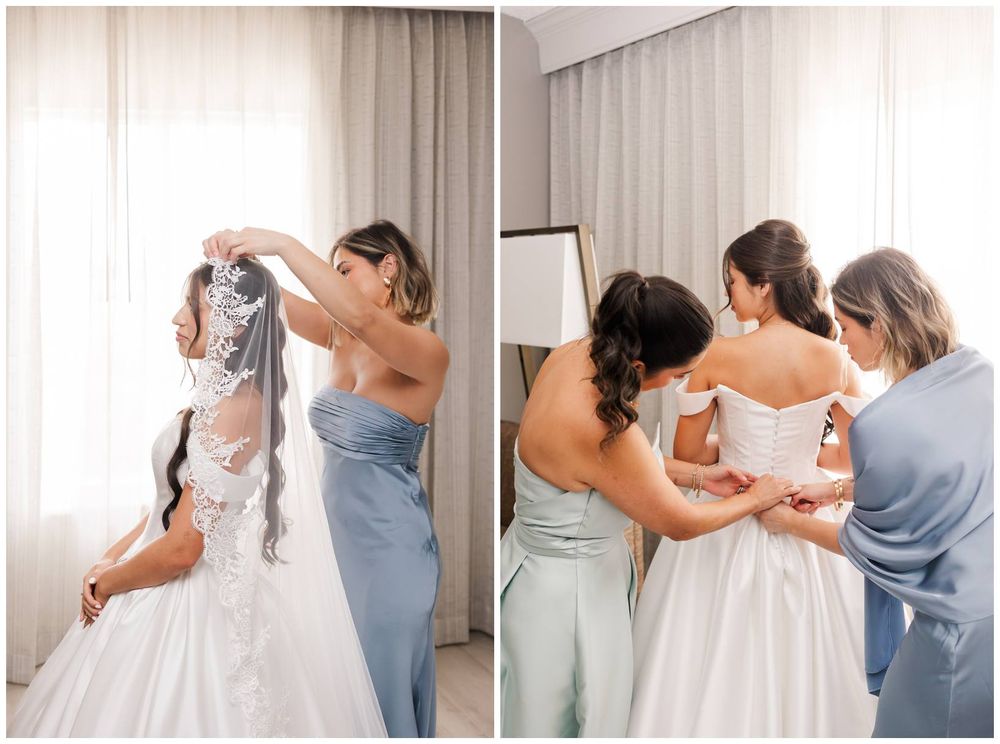 Bridesmaids in blue dresses helping bride into wedding gown and veil in bright hotel room.