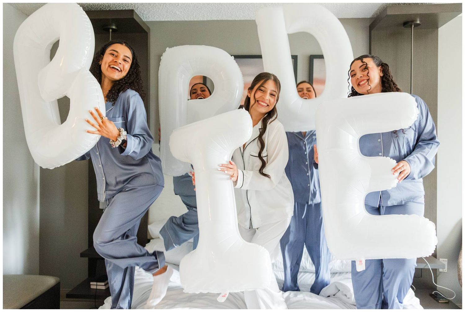 Four bridesmaids in satin pajamas hold giant white BRIDE balloon letters while standing on a bed.