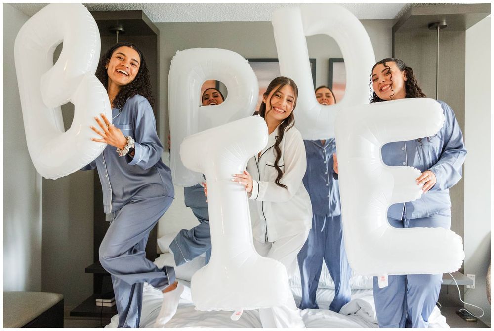 Four bridesmaids in satin pajamas hold giant white BRIDE balloon letters while standing on a bed.