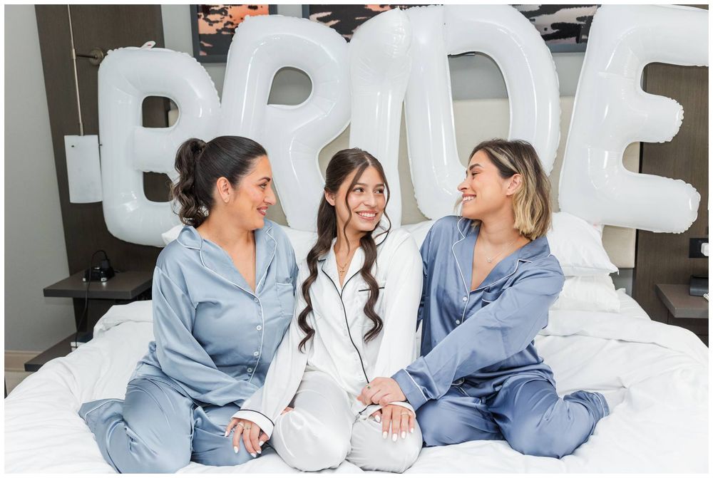 Three women in satin pajamas sit on a bed with white BRIDE balloons, celebrating a bridal party moment.