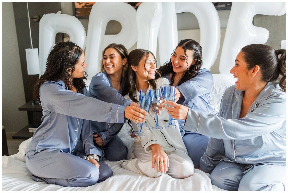 Bridesmaids in matching blue satin pajamas toasting champagne on a bed with a BRIDE balloon backdrop.