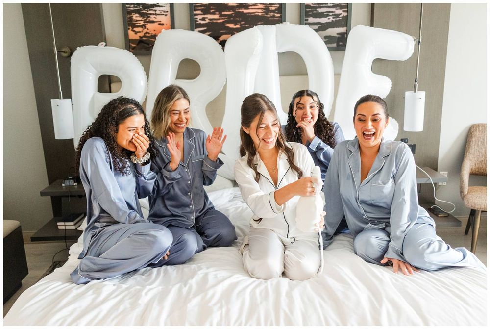 Five women in matching satin pajamas sit on a bed laughing, with large balloon letters spelling BRIDE behind them.