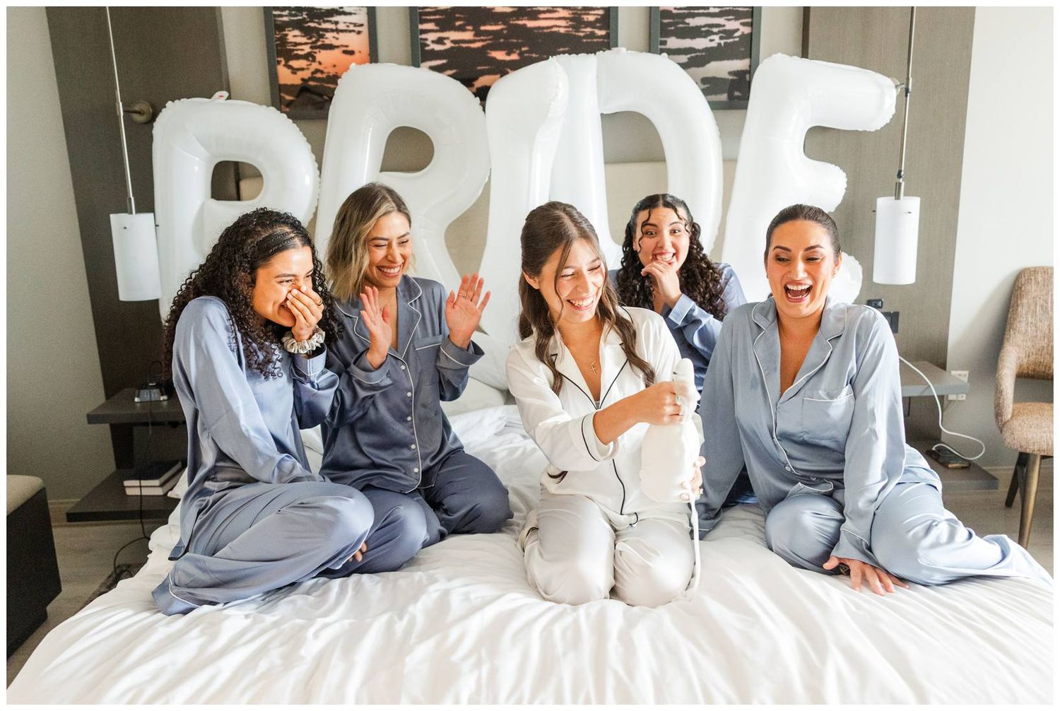 Five women in matching satin pajamas sit on a bed laughing, with large balloon letters spelling BRIDE behind them.