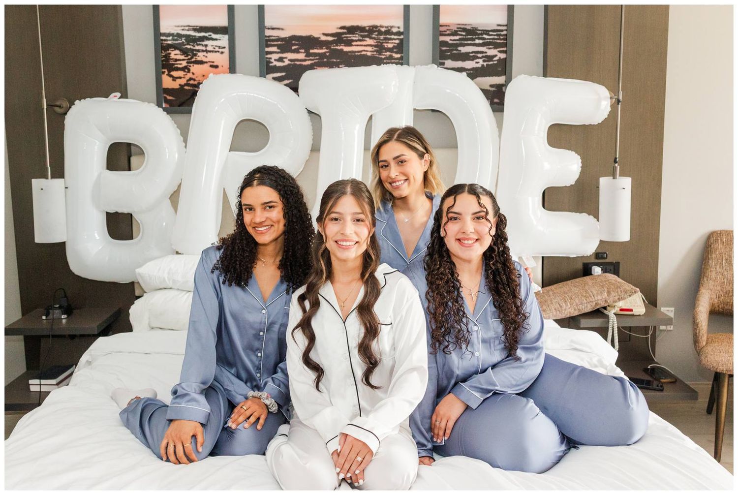 Four women in blue and white satin pajamas sit on a bed in front of large white BRIDE balloon letters.