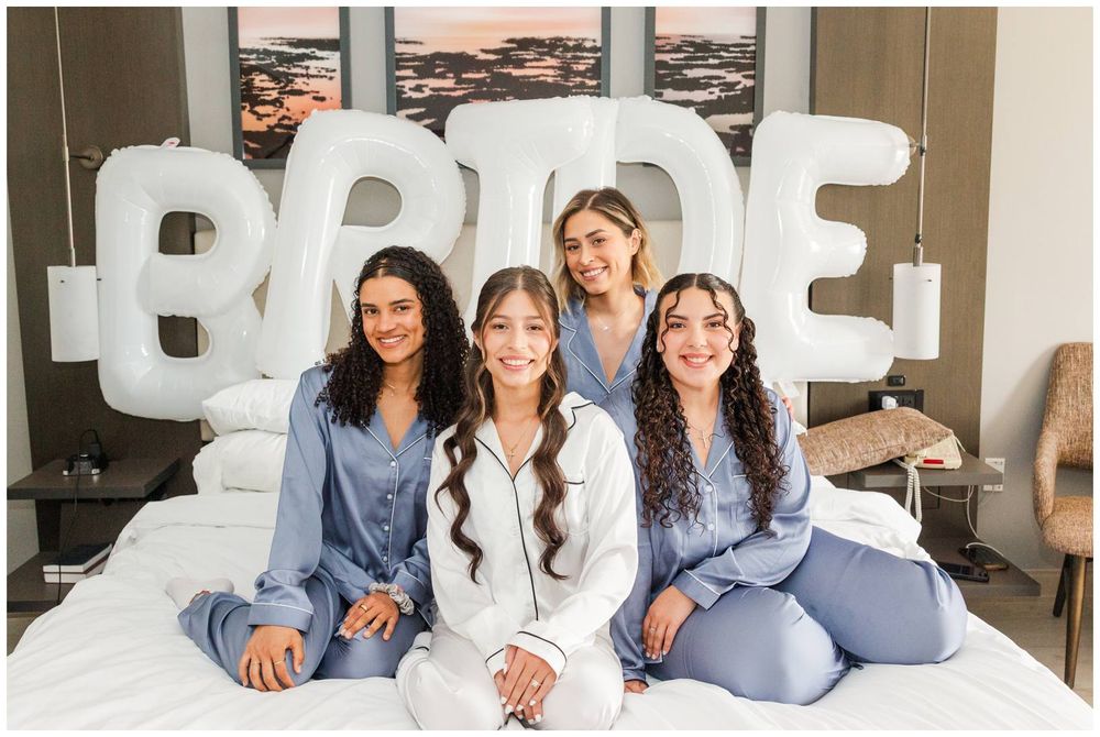 Four women in blue and white satin pajamas sit on a bed in front of large white BRIDE balloon letters.