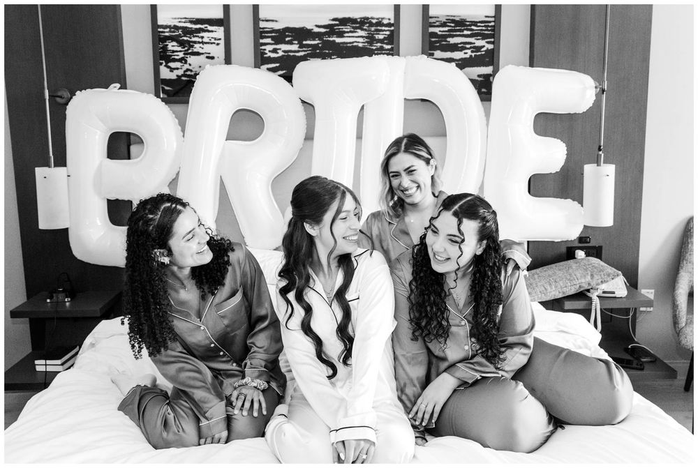 Bride and bridesmaids laughing on bed with large BRIDE balloon letters in black and white wedding photo.